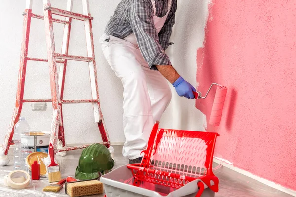 Caucasian house painter worker in white work overalls, with the roller painting the wall with colorful painting. Construction industry.