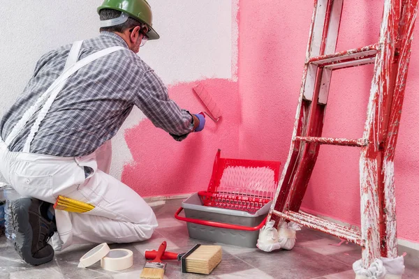 Caucasian house painter worker in white work overalls, with the roller he paints the wall with the colored painting of pink. Construction industry. Work safety. Bottom view.