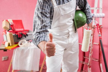 Beyaz iş tulumlu beyaz boyacı, baş parmağı havada 