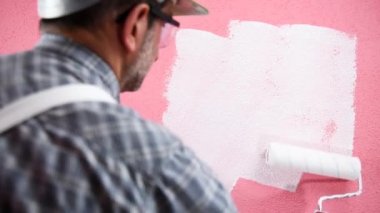 Beyaz tulumlu, kasklı ve koruyucu gözlüklü kafkas ev ressamı. Rulo ile beyaz boya ile pembe duvar boyalar. İnşaat sektörü. Görüntü.