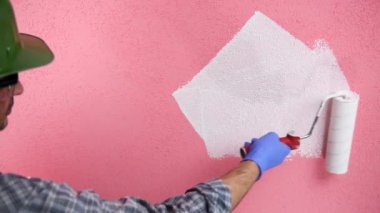 Beyaz tulumlu, kasklı ve koruyucu gözlüklü kafkas ev ressamı. Rulo ile beyaz boya ile pembe duvar boyalar. İnşaat sektörü. Görüntü.