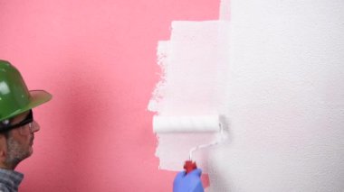 Beyaz tulumlu, kasklı ve koruyucu gözlüklü kafkas ev ressamı. Rulo ile beyaz boya ile pembe duvar boyalar. İnşaat sektörü. Görüntü.