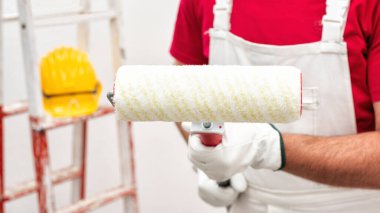 Beyaz iş tulumlu, beyaz tenli boyacı ve koruyucu eldivenleriyle elinde boya için silindiri tutuyor. İnşaat endüstrisi. Çalışma araçları.