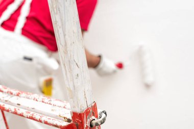 Beyaz tulumlu, koruyucu eldivenli, beyaz boyalı bir ev boyacısı. Duvarı silindir kullanarak beyaz boyayla boyuyor. İnşaat endüstrisi. İş güvenliği.