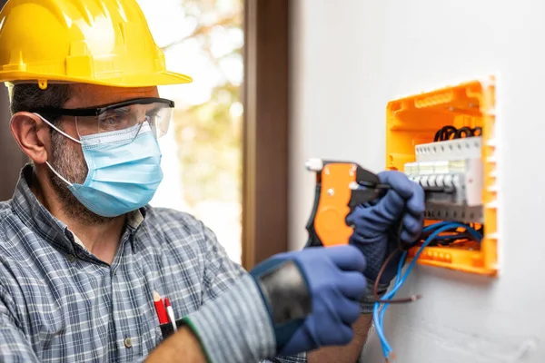 Electrician at work on an electrical panel protected by helmet, safety goggles and gloves; wear the surgical mask to prevent the spread of Coronavirus. Construction industry. Covid-19 Prevention.