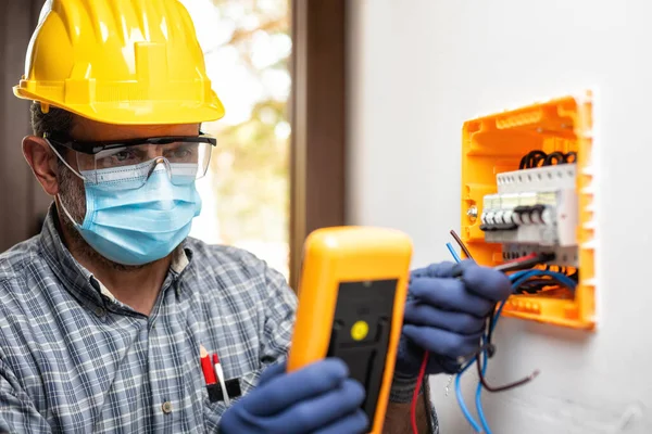 Electrician at work on an electrical panel protected by helmet, safety goggles and gloves; wear the surgical mask to prevent the spread of Coronavirus. Construction industry. Covid-19 Prevention.
