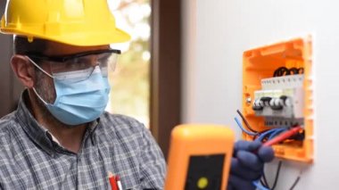 Kask, güvenlik gözlükleri ve eldivenlerle korunan bir elektrik paneli üzerinde çalışan elektrikçi Coronavirus 'un yayılmasını önlemek için cerrahi maske takıyor. İnşaat endüstrisi. Covid-19 Salgın Önleme. Görüntü.
