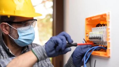 Kask, güvenlik gözlükleri ve eldivenlerle korunan bir elektrik paneli üzerinde çalışan elektrikçi Coronavirus 'un yayılmasını önlemek için cerrahi maske takıyor. İnşaat endüstrisi. Covid-19 Salgın Önleme. Görüntü.