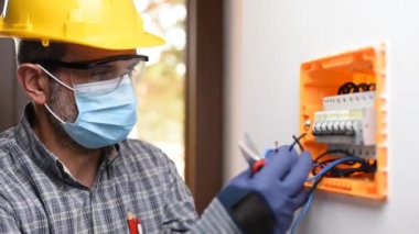 Kask, güvenlik gözlükleri ve eldivenlerle korunan bir elektrik paneli üzerinde çalışan elektrikçi Coronavirus 'un yayılmasını önlemek için cerrahi maske takıyor. İnşaat endüstrisi. Covid-19 Salgın Önleme. Görüntü.