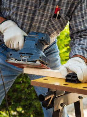 Yakın plan. Elleri elektrikli zımpara makinesi ile korunan marangoz ahşap bir tahtayı yumuşatıyor. İnşaat endüstrisi. İş güvenliği.