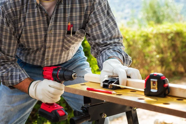 Close-up. Carpenter with his hands protected by gloves with the electric drill, drill a wooden board. Construction industry. Work safety.