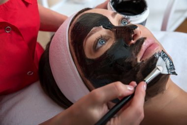 Güzellik salonunda, tedavi edici siyah çamurun yüzü için maske takan genç ve güzel bir kadın. Spa, cilt bakımı, cilt temizleme ve nemlendirici. Sağlık ve rahatlama..