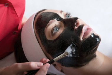 Güzellik salonunda, tedavi edici siyah çamurun yüzü için maske takan genç ve güzel bir kadın. Spa, cilt bakımı, cilt temizleme ve nemlendirici. Sağlık ve rahatlama..