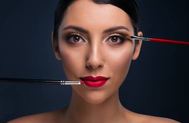 Parlak makyajlı ve iki kozmetik fırçası olan güzel bir manken. Renkli dumanlı gözleri ve kırmızı dudakları olan genç bir kadın. Uzun takma kirpikler. Profesyonel stil. 