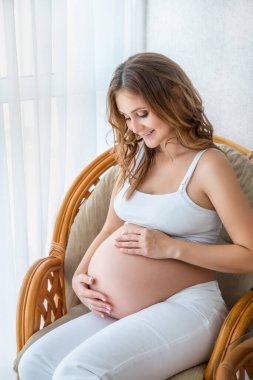 Hamile kadın pencerenin önünde oturur ve karnına dokunur. Hamilelik ve aile. Yaşam biçimi.