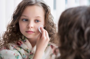 Güzel küçük bir kızın portresi ev banyosundaki aynanın önünde cilt temizleme işleminden zevk alıyor. Güzel çocuk yüzünü pamukla ve nemlendirici losyonla temizliyor. Gençlik ve çocuk cilt sorunları, hijyen, temiz ve taze genç