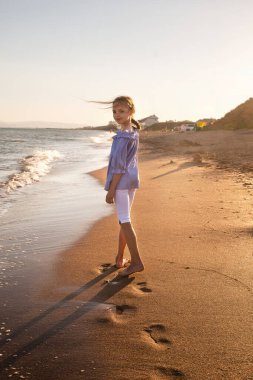 Gün batımında deniz dalgalarında yürüyen güzel küçük bir kızın portresi. Çocuk ve tatil, turistik bir yer..