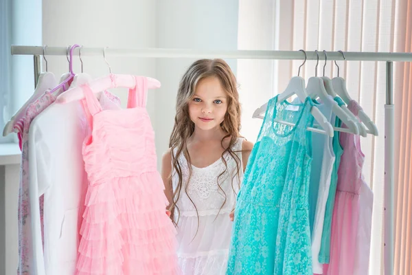 Little child girl choosing her clothes. Kid thinking what to choose to wear in front of many choices of dresses on hangers. Sales, buy and sell concepts.