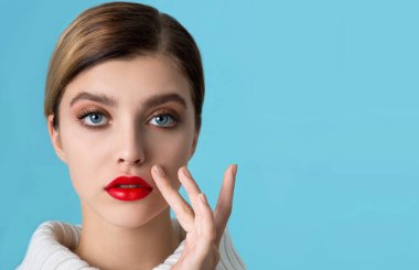 Genç ve güzel bir kadının portresi, mükemmel bir cilt, beyaz kürklü kırmızı dudaklar. Açık mavi arka planda parlak kırmızı makyajlı ve parıltılı manikürlü bir model. Kış mevsimi güzellik konsepti. Yakın çekim, kopyalanma alanı.