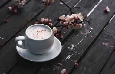 Bir fincan sıcak kahve eski kırsal siyah ahşap masa üstünde bahar çiçekleriyle. Beyaz sütlü kapuçino ve sezonluk arka planda tarçın..