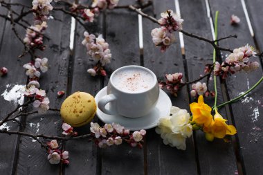 Bir fincan sıcak kahve, badem kurabiyesi ve bahar çiçeği, eski kırsal siyah ahşap masa üstü. Beyaz sütlü kapuçino ve sezonluk arka planda tarçın..