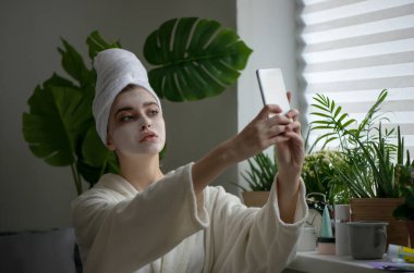 Yüzünde krem çamur maskesi olan genç bir kadın telefonunda selfie çekiyor. Güzellik blogcusu, SPA prosedürleri sırasında akıllı telefondan baktığını gösteriyor. Spa ve Sağlık. Kapat, seçili odak.