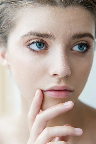 Beautiful young woman with clean perfect skin and water moisture drops touching face. Portrait of beauty model with natural nude makeup moisturizing her skin by lotion. Spa, skincare and wellness. Close up, background, copy space.