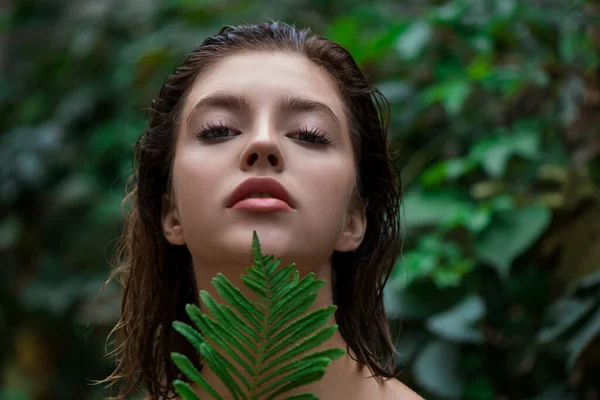 Beautiful young woman with perfect skin and natural make up posing front of plant tropical green leaves background with fern. Teen model with wet hair care of her face and body. SPA, wellness, bodycare and skincare.  Close up, selective focus.