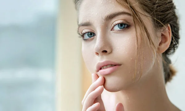 Beautiful young woman with clean perfect skin and water moisture drops touching face. Portrait of beauty model with natural nude makeup moisturizing her skin by lotion. Spa, skincare and wellness. Close up, background, copy space.