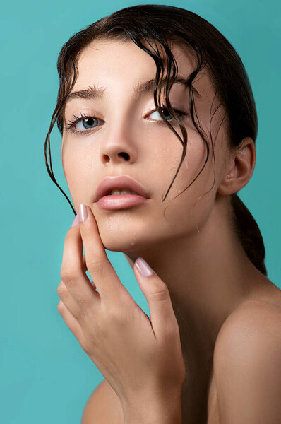 Beautiful young woman with clean perfect skin and water moisture drops touching face. Portrait of beauty model with natural nude makeup moisturizing her skin by lotion. Spa, skincare and wellness. Close up, background, copy space.