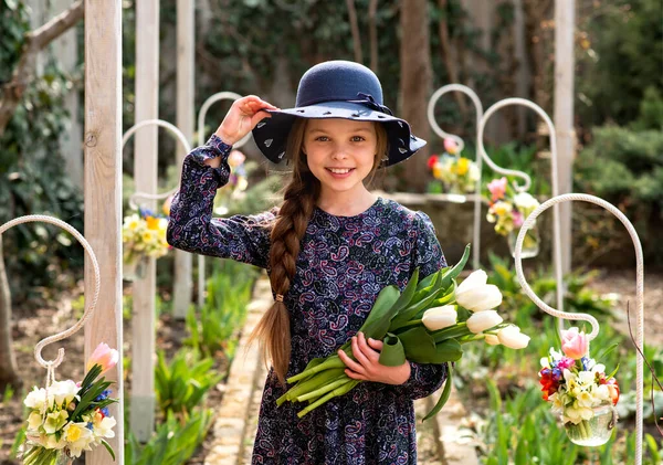 手の屋外でチューリップ花束と帽子のかわいい笑顔の女の子 春のイースターの季節の肖像画 田舎を背景に春の花を持つ子供 — ストック写真