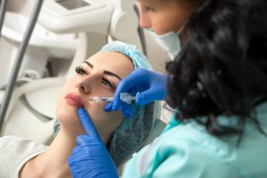 Kozmetik uzmanının elleri yüzüne ve dudaklarına iğne yapıyor. Genç bir kadın güzellik salonunda yüz aşısı yaptırıyor. Yaşlanma, gençleşme ve hidrasyon prosedürleriyle yüzleş. Estetik kozmetoloji. Kapat..
