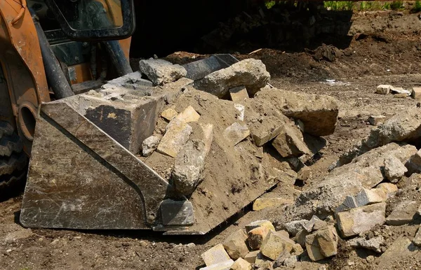 The bucket of an excavating machine is loaded with gravel and chunks of broken concrete.