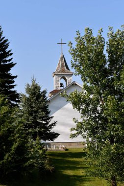 Bir çan kulesi ve eski beyaz kilise binası arkası çam ağaçları görülebilir. 