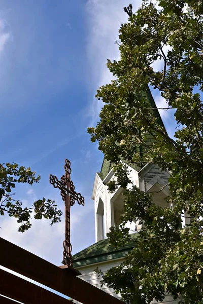 A metal cross on a beam is flanked by the partially hidden steeple on a white church.