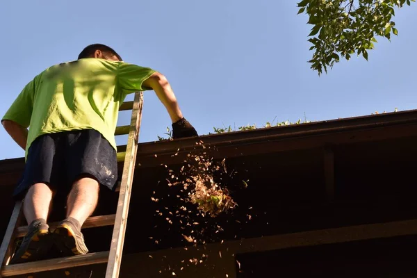 An unidentified man stands on an extension ladder cleaning the eaves of a two story house