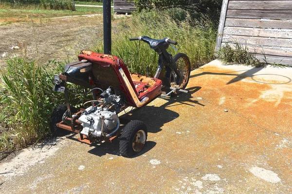 An old three wheeled go-cart in disrepair is parked in front of an abandoned garage.