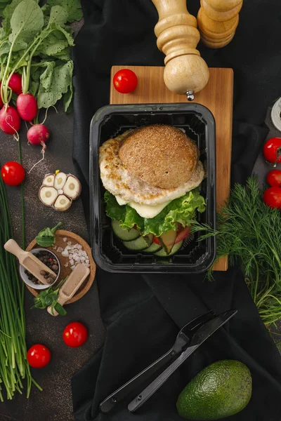 Food composition of burger with chicken cutlet, lettuce and vegetable salad in black tray on table with vegetables and kitchenware