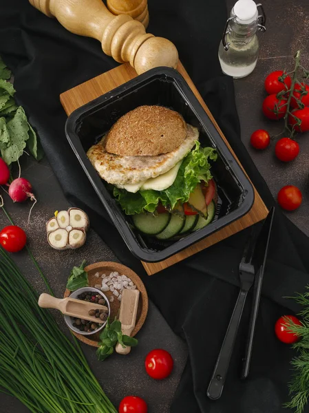 Food composition of burger with chicken cutlet, lettuce and vegetable salad in black tray on table with vegetables and kitchenware