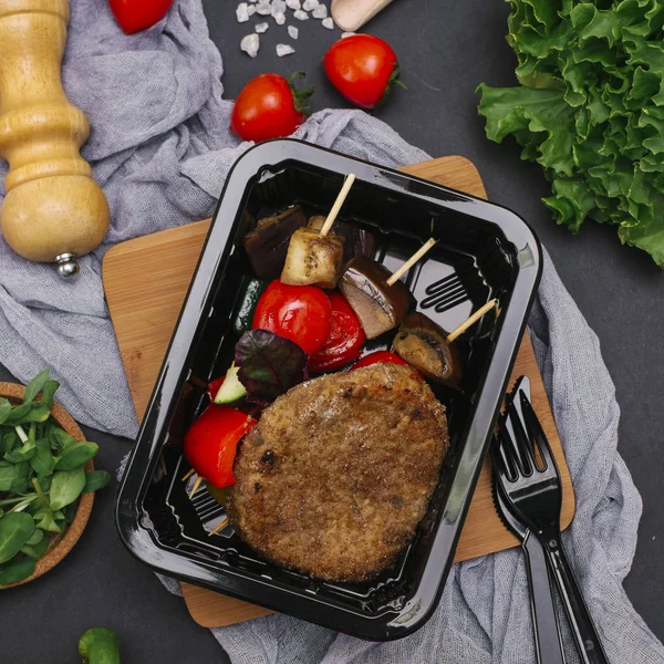 Breaded meat cutlet with grilled vegetables on skewers in black tray with fork and knife