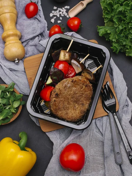 Breaded meat cutlet with grilled vegetables on skewers in black tray with fork and knife