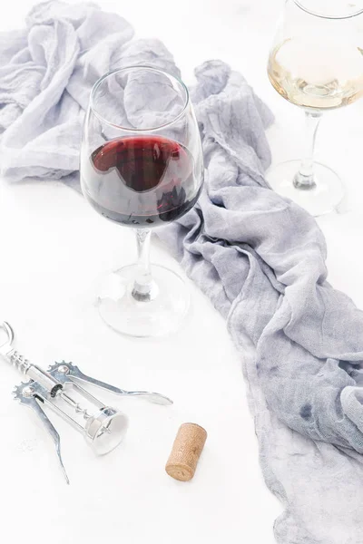 Glasses of red and white wine on white concrete surface with cork, corkscrew and linen cloth