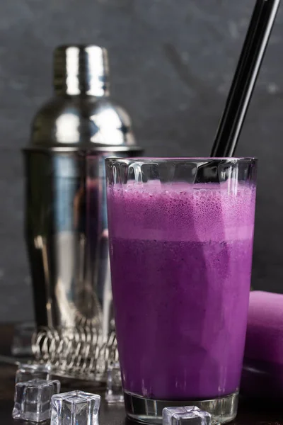 Glass of purple drink and cocktail shaker with ice cubes on table, still life