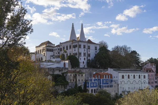 Wide view of the Sintra National Palace in Portugal with its iconic twin chimneys, surrounded by colorful houses and lush vegetation against a blue sky.