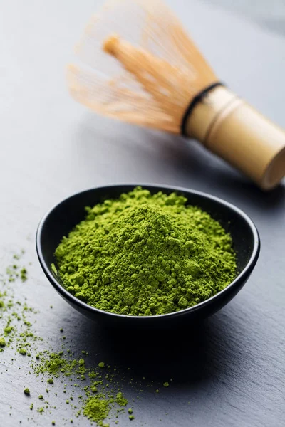 Matcha, green tea powder in black bowl with bamboo whisk on slate background.