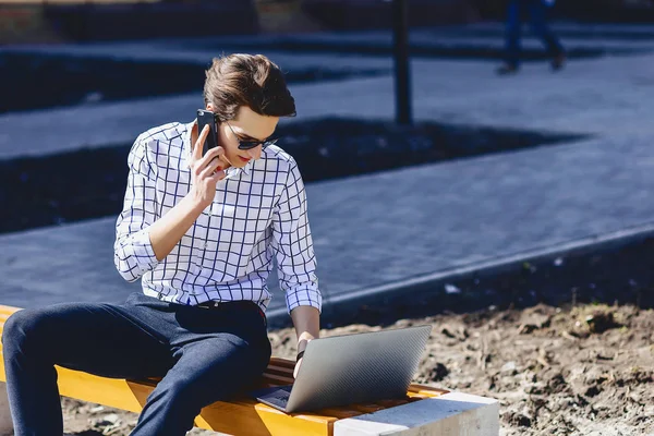 Young stylish man with phone work on laptop at park on sunny day
