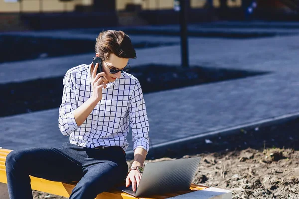 Young stylish man with phone work on laptop at park on sunny day