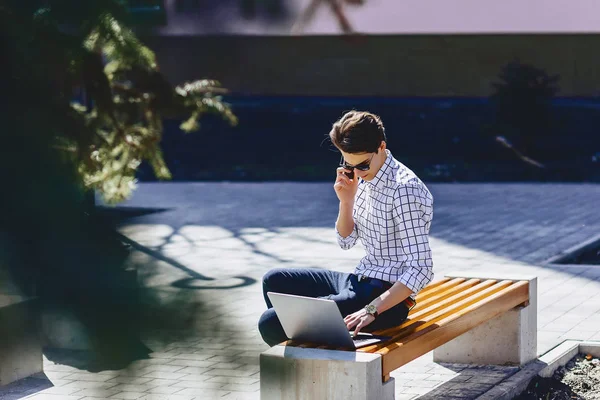 Young stylish man with phone work on laptop at park on sunny day