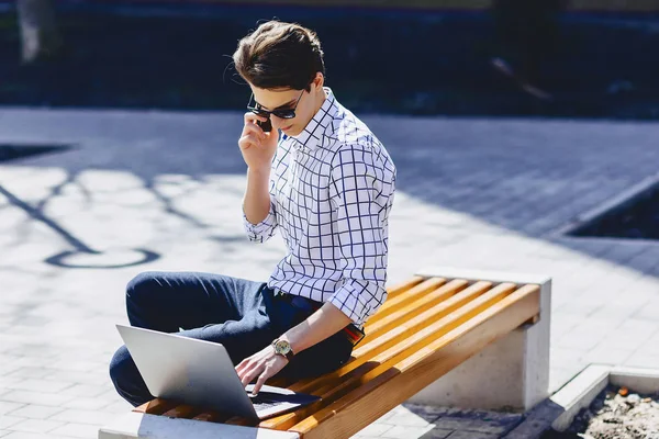 Young stylish man with phone work on laptop at park on sunny day