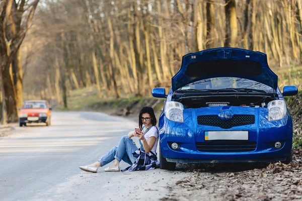 girl is sitting by car with an open hood and speaks by phone on road alone